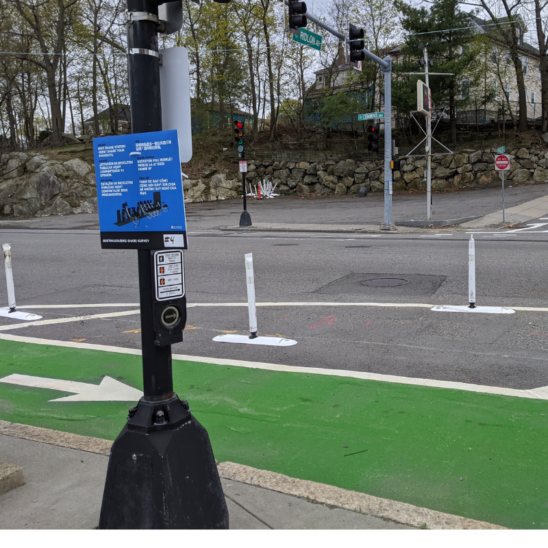 A blue sign located on the pole of a pedestrian signal.  It is next to a green bike lane with white flexposts and a white arrow.  The sign has information about potential Bluebikes station locations in the area