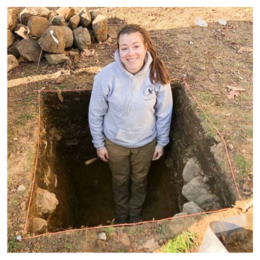 A smiling woman with dark hair wearing a grey sweatshirt. She is standing in an archaeological excavation unit with a pile of rocks in the background.