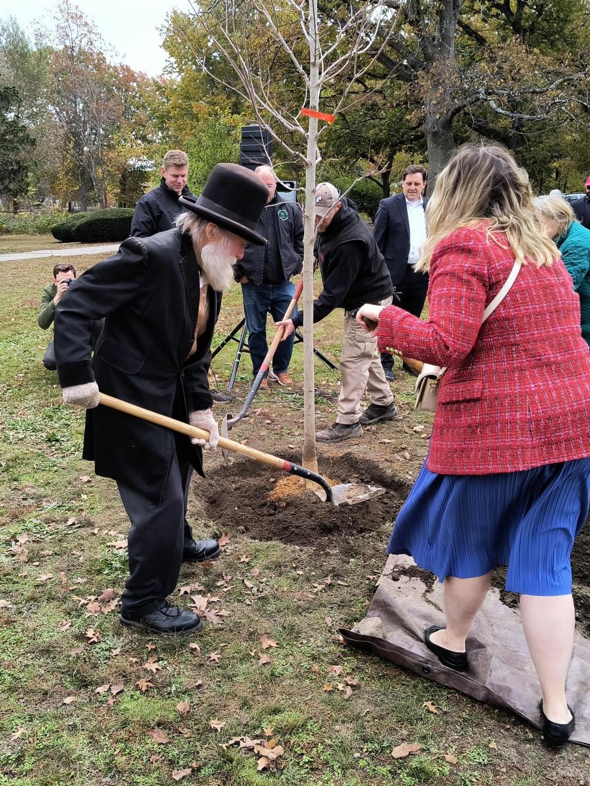 A local personality dressed to embody Frederick Law Olmstead shovels dirt onto the newly planted tree with a woman next to him.