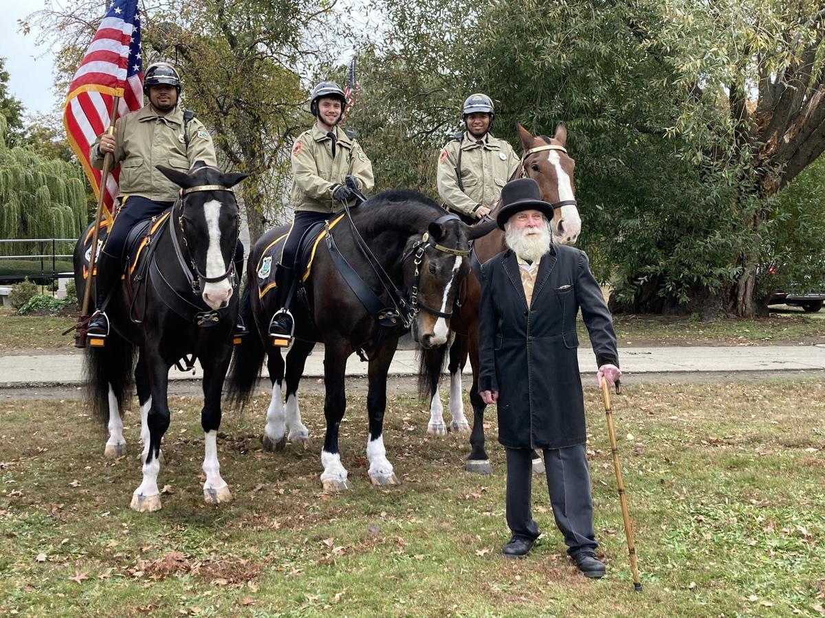 A local personality dressed to embody Frederick Law Olmstead poses in front of three mounted Parks rangers and a flag.