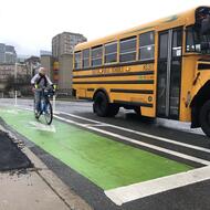 An adult bicycles toward us in a green bike. A yellow school bus is in the adjacent travel lane.
