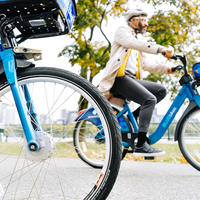 A Bluebikes rider is riding on a path. A second Bluebikes bike is in the foreground, with a focus on the front wheel.