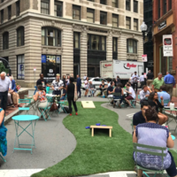 A photo showing a block party on the street featuring people gathered and socializing around movable street furniture, artificial turf, and lawn games
