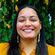 A picture of a person in front of a tree. The person has long, dark hair, and is wearing a mustard-color shirt.