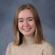 Headshot of Morgan Clark, a white woman with blonde shoulder-length hair wearing a cream-colored turtleneck, smiling at the camera in front of a blue-gray background