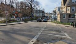 A photograph taken from the southeast corner of the intersection of Claybourne Street and Tonawanda Street, looking north up Claybourne Street. A playground in Mother's Rest Park can be seen just beyond the intersection. Two people are walking towards a staircase to enter the park.