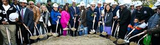Mayor Michelle Wu with a large group of community leaders, officials, and residents wearing white hard hats stand shoulder to shoulder outdoors, holding shovels filled with sand for a ceremonial groundbreaking. A modern building is visible in the background, and the group includes adults and a child, all facing the camera and smiling.